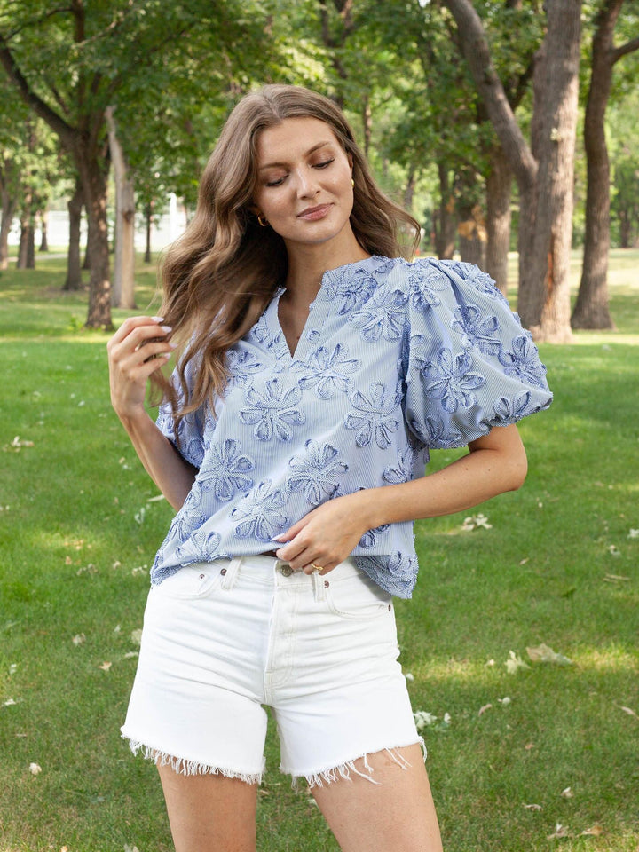 A woman standing outdoors in a park wearing a light blue floral puff-sleeve blouse and white denim shorts.