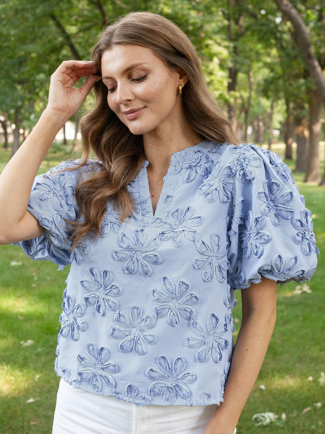 A woman wearing a light blue puff-sleeve blouse with tonal floral embroidery, standing outdoors in a park with green trees in the background.