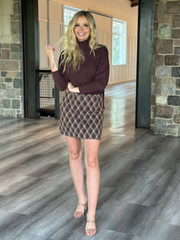 A woman modeling a dark brown mock neck long-sleeve top and a brown and beige plaid mini skirt, standing in a modern indoor hallway with stone columns and wood floors.