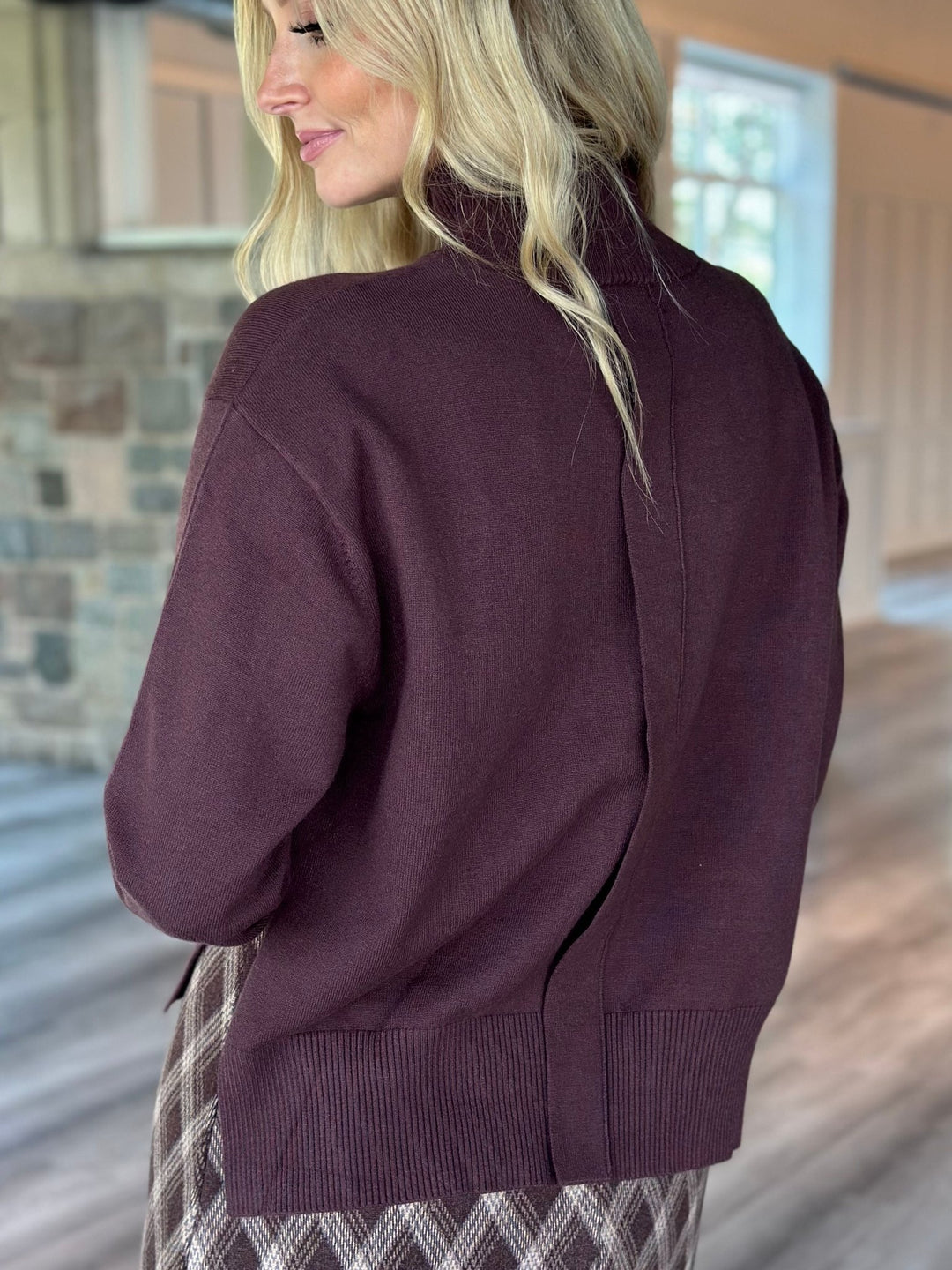 A woman shown from the back wearing a dark plum purple knit cardigan sweater, paired with a plaid skirt, in a rustic indoor setting with a stone wall and wood floor.