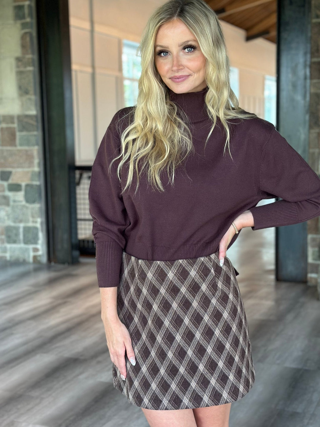A woman modeling a dark plum turtleneck sweater paired with a brown and beige plaid mini skirt, standing in a modern indoor hallway with stone columns and wood floors.