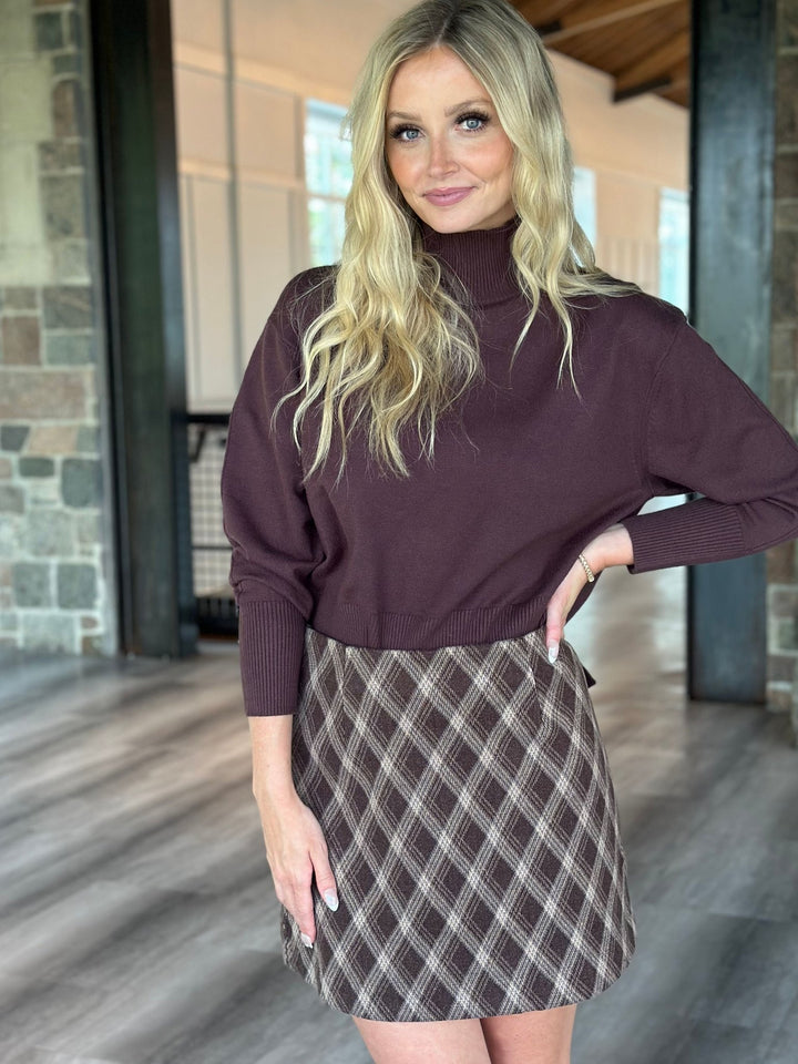 A woman modeling a dark plum turtleneck sweater paired with a brown and beige plaid mini skirt, standing in a modern indoor hallway with stone columns and wood floors.