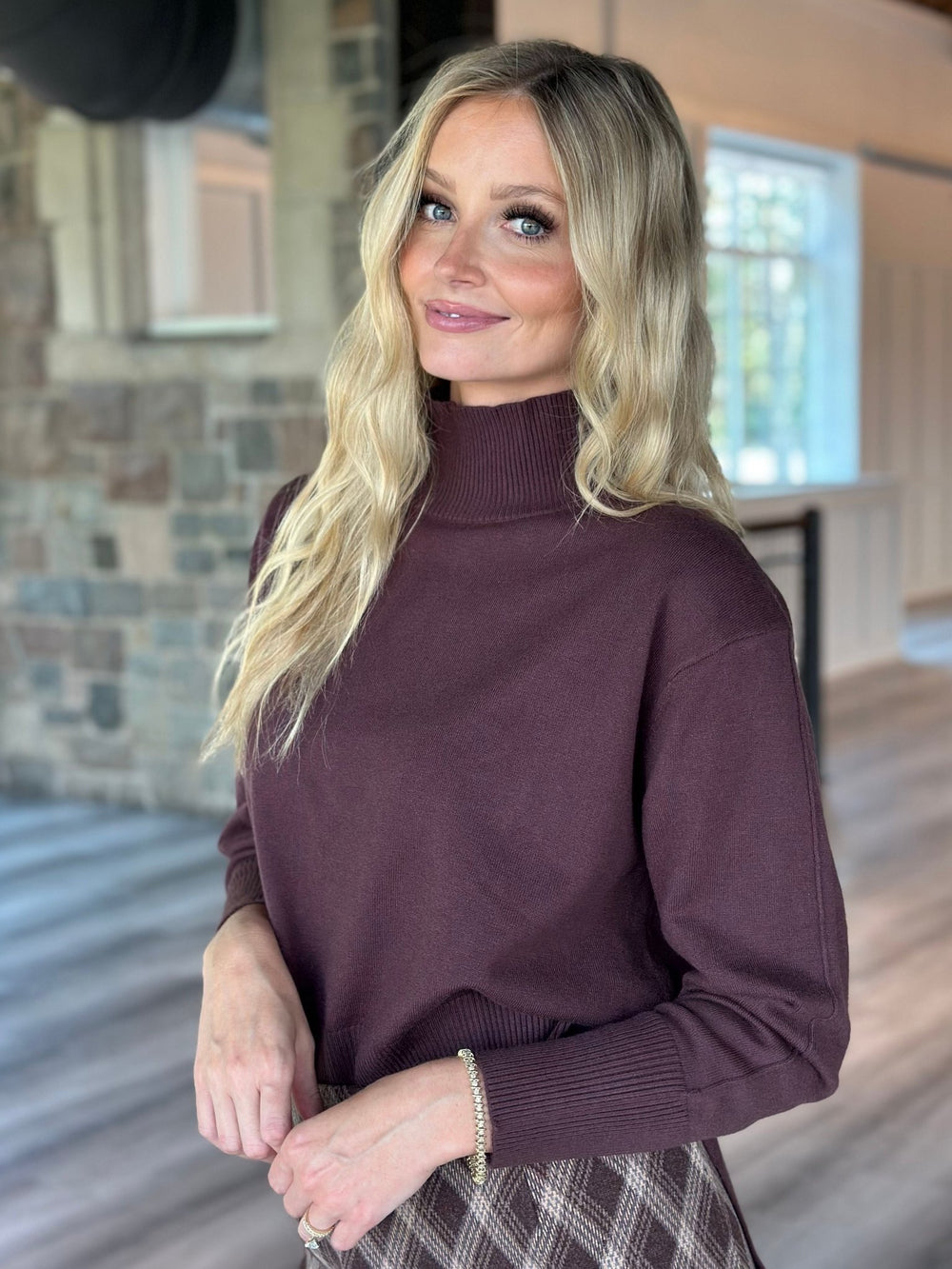 A blonde woman wearing a long-sleeve ribbed mock neck sweater in a deep plum purple, standing in a home interior with wood floors and a stone wall.