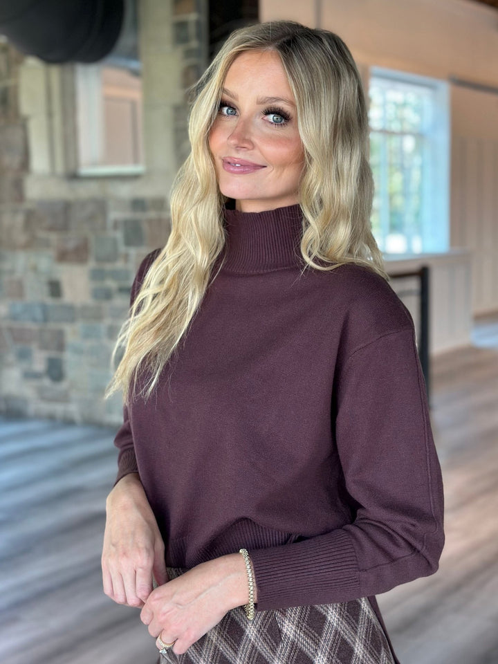 A blonde woman wearing a long-sleeve ribbed mock neck sweater in a deep plum purple, standing in a home interior with wood floors and a stone wall.
