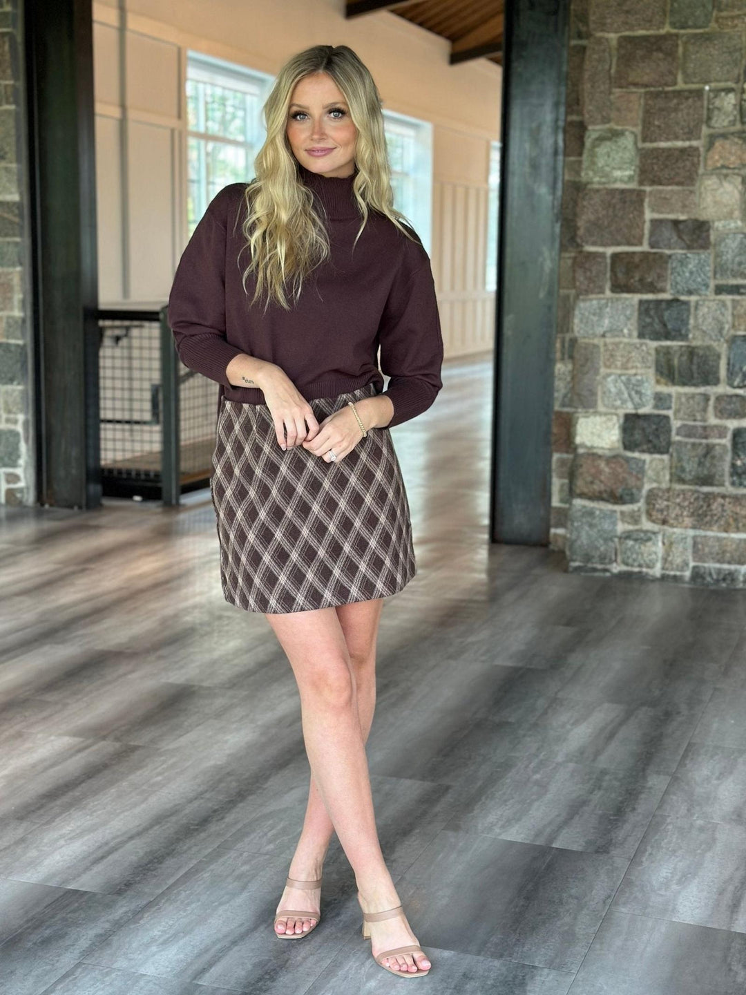 A woman modeling a dark plum long-sleeve mock neck top and a brown and gray plaid mini skirt, standing in a modern indoor hallway with wood floors and a stone pillar.
