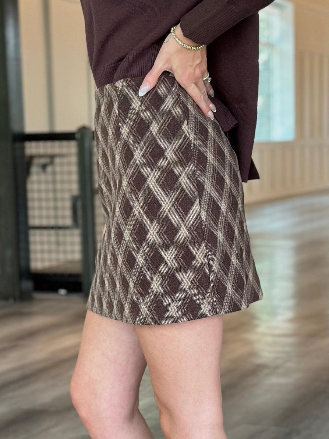 A person wearing a brown and beige diamond-patterned mini skirt with a dark brown ribbed top, standing in an indoor space with a wooden floor.