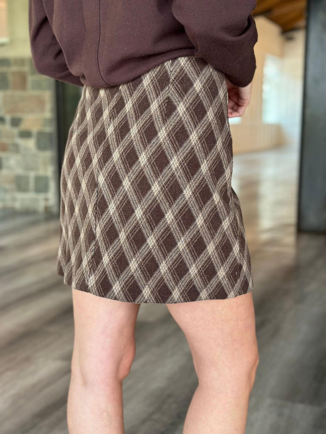 A person wearing a brown and beige plaid mini skirt with a dark brown top, standing in a modern indoor space with a blurred stone and wood background.
