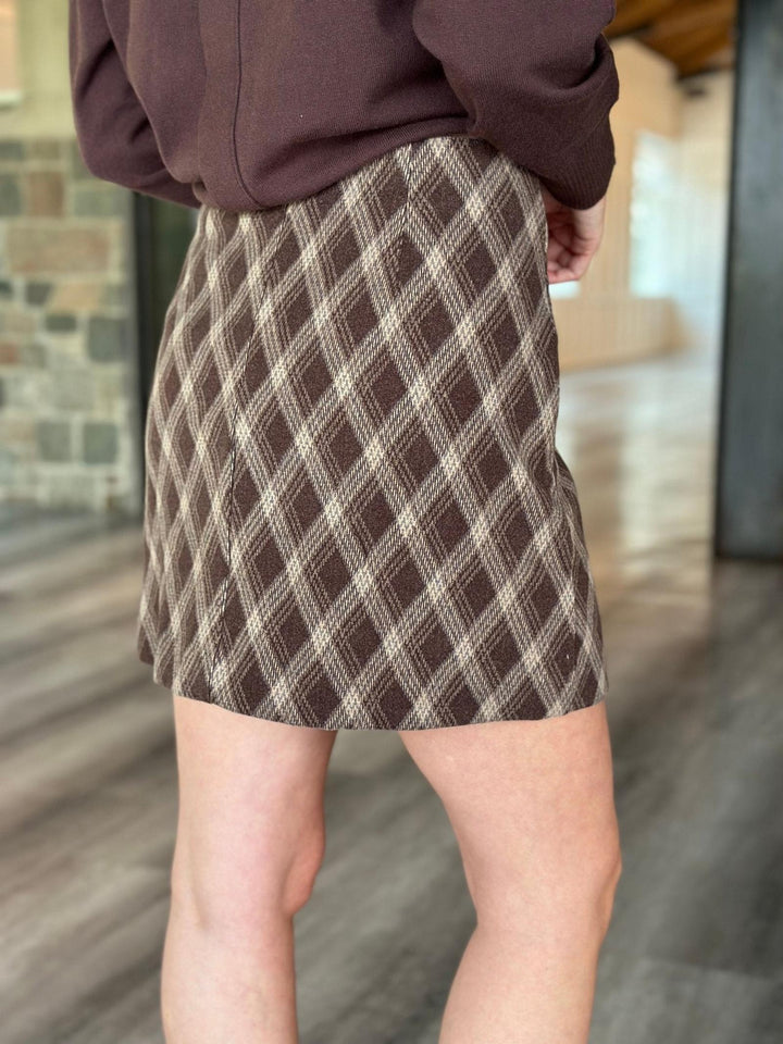 A person wearing a brown and beige plaid mini skirt with a dark brown top, standing in a modern indoor space with a blurred stone and wood background.