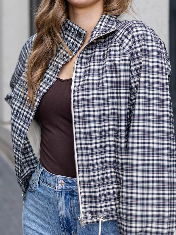 A woman wearing a navy and white plaid zip-up jacket over a dark brown tank top and blue jeans, shown in a close-up outdoor shot.