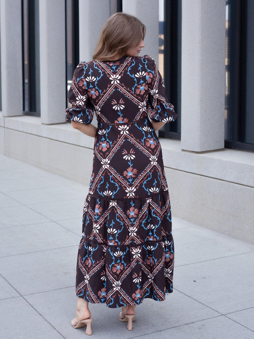 A woman viewed from behind wearing a long-sleeved tiered maxi dress with a dark brown base and a colorful geometric and floral pattern in blue, pink, and white. She is standing outdoors on a light gray tiled walkway in front of a modern building with gray columns.
