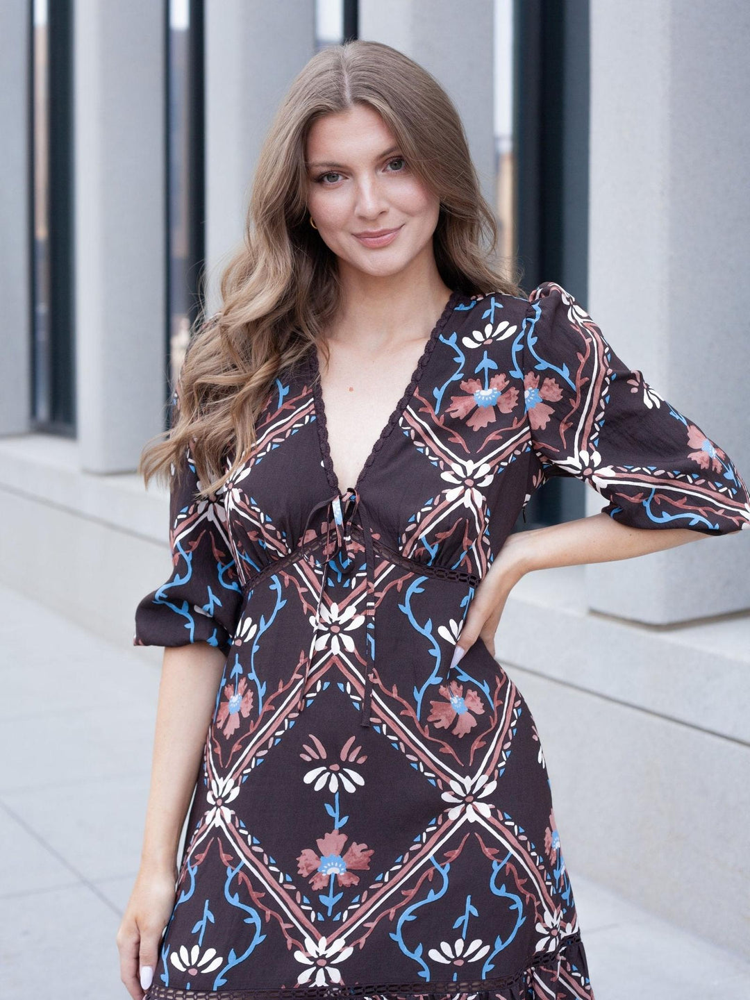 A woman modeling a dark brown bohemian-style dress with a blue, pink, and white floral geometric pattern, standing outdoors against a modern gray building background.