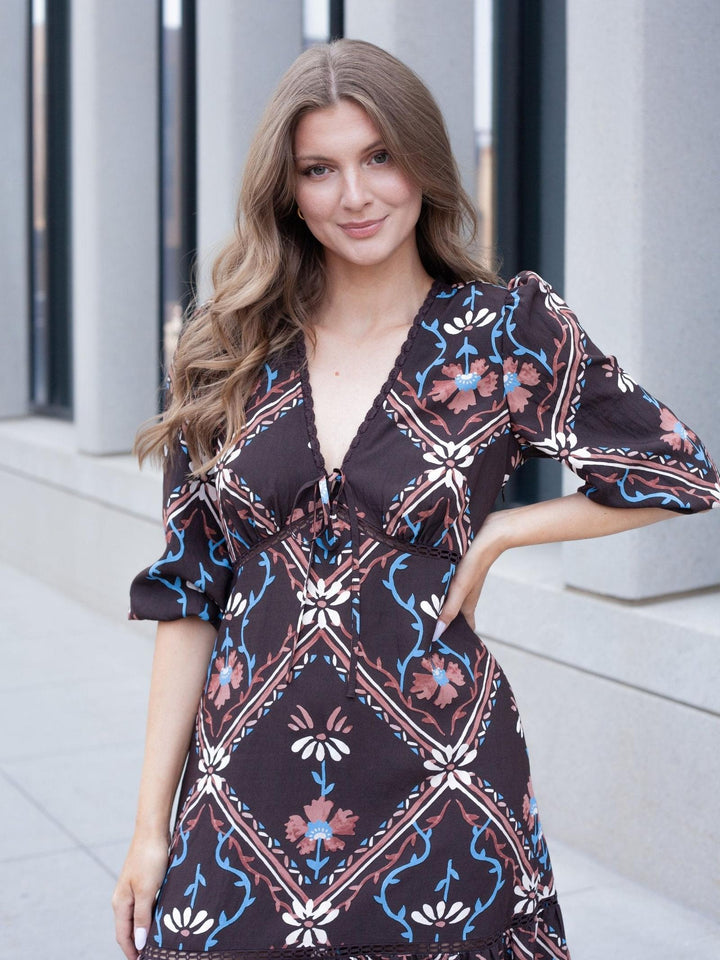 A woman modeling a dark brown bohemian-style dress with a blue, pink, and white floral geometric pattern, standing outdoors against a modern gray building background.