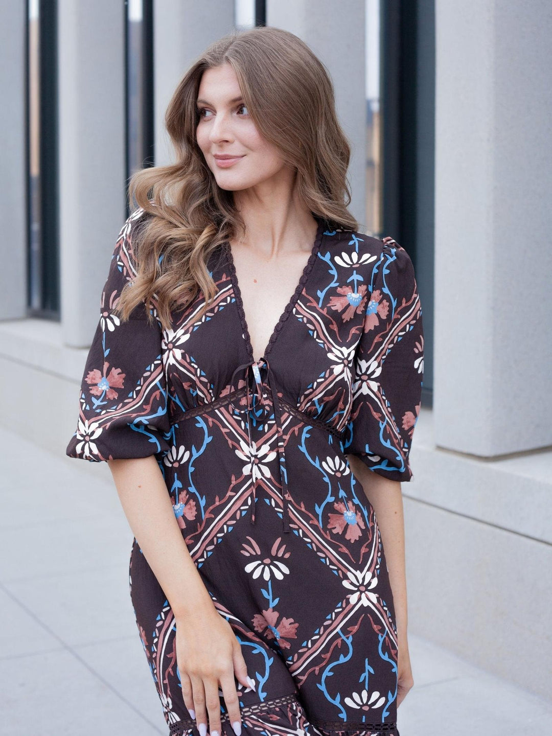 A woman wearing a dark brown jumpsuit with a colorful floral and geometric pattern, standing outdoors in front of a modern concrete building.