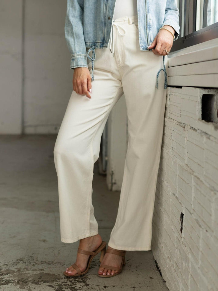 Person wearing white pants and a denim jacket leaning against a wall.