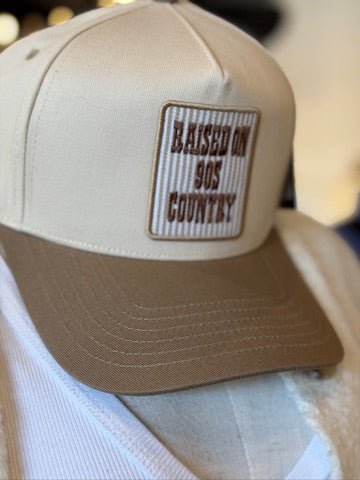 A beige and brown trucker-style cap with a rectangular front patch reading 'RAISED ON 90S COUNTRY' in brown text on a white-and-brown striped background.