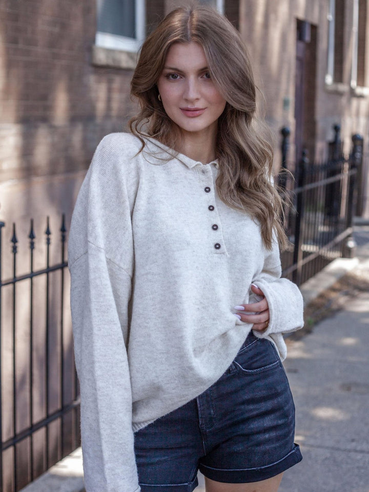 A woman wearing a light oatmeal-colored knit henley sweater and dark denim shorts, standing outdoors on a sidewalk in front of a brick building and black metal fence.