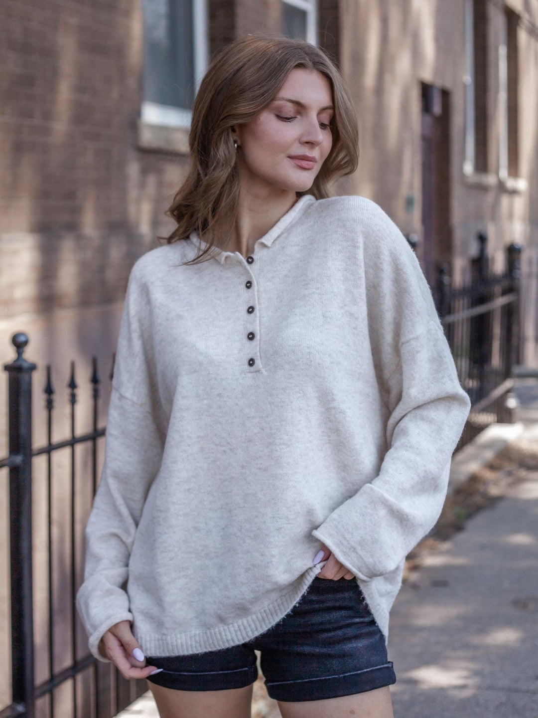 A woman wearing an oversized light beige knit henley sweater with a buttoned collar, paired with dark denim shorts, standing on a city sidewalk.