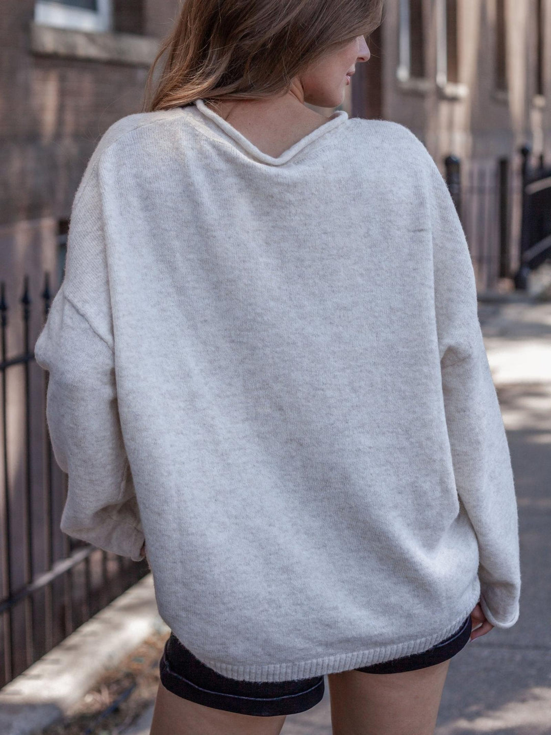 A woman wearing an oversized light gray knit sweater, shown from the back in an outdoor urban setting.