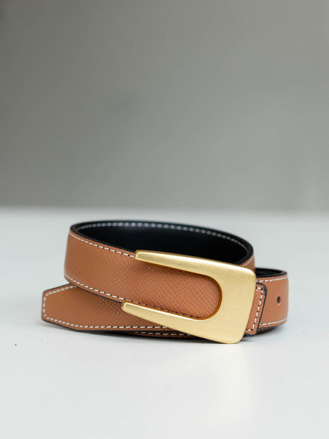 Tan leather reversible belt with angular asymmetrical gold buckle and white stitching.