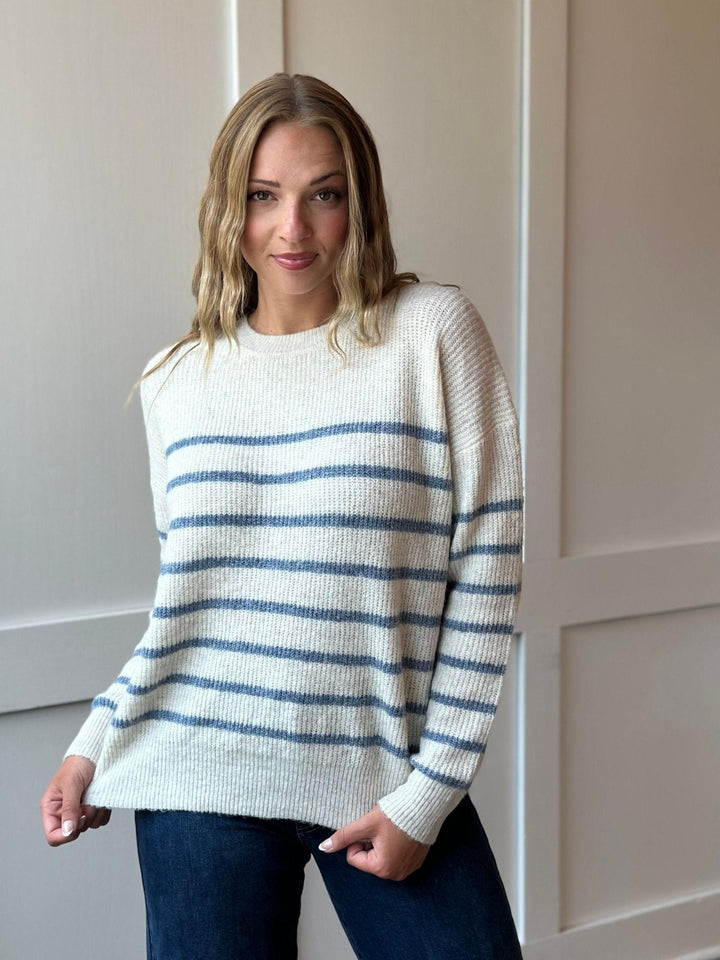 A woman wearing a cream-colored knit sweater with horizontal blue stripes, paired with dark blue jeans, standing against a beige paneled wall.