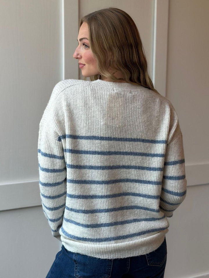 A woman shown from the back wearing a cream-colored ribbed knit sweater with horizontal blue stripes, paired with dark blue jeans, standing against a neutral indoor wall.