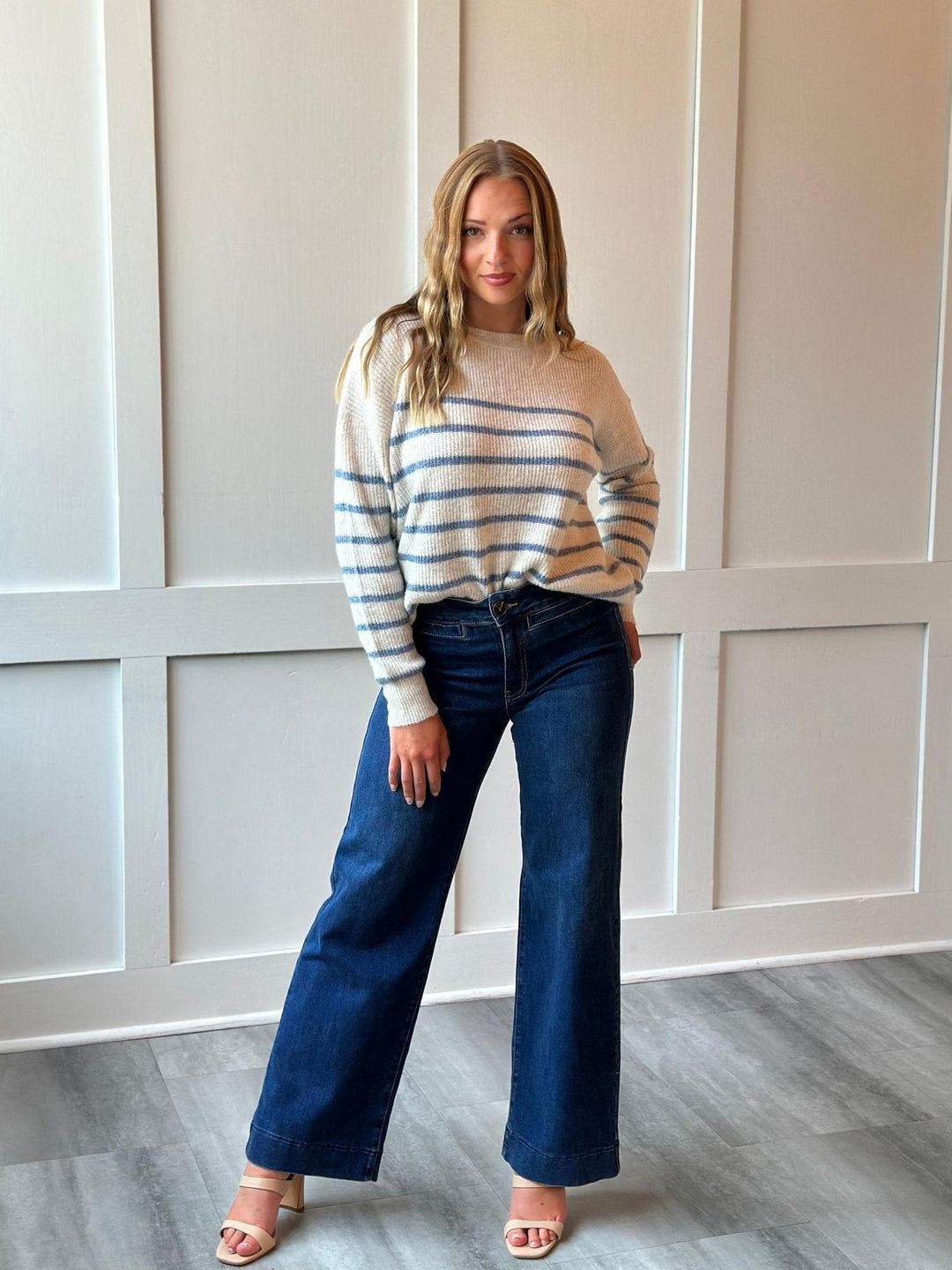 A woman modeling a beige and navy blue striped knit sweater paired with dark blue wide-leg jeans and nude heeled sandals, standing in front of a white paneled wall.