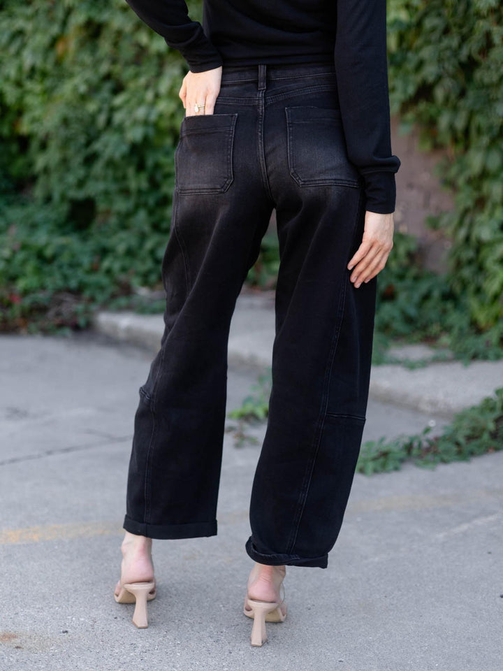 A person shown from the back wearing black wide-leg cropped jeans, a black long-sleeve top, and beige heeled sandals on an outdoor paved path with green foliage in the background.
