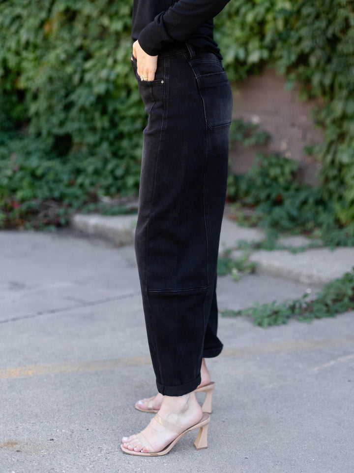 A person wearing black wide-leg cropped jeans with a relaxed, baggy fit, paired with a black long-sleeve top and clear high-heel mules, standing outdoors on a paved path with green foliage in the background.