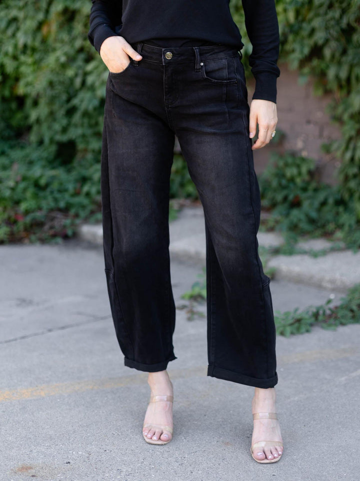 A person wearing black wide-leg cropped jeans with side pockets, paired with a black long-sleeve top and clear high-heel sandals, standing outdoors on a paved surface with green foliage in the background.