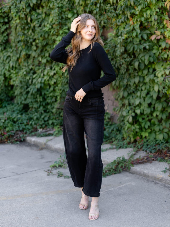 A woman wearing a black long-sleeve top and black wide-leg jeans stands on a concrete path in front of a green ivy-covered wall.