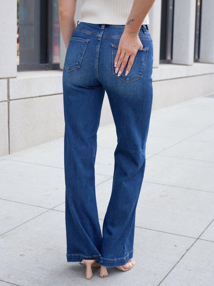 A person shown from the back wearing blue wide-leg jeans, standing on a light stone sidewalk in front of a building.