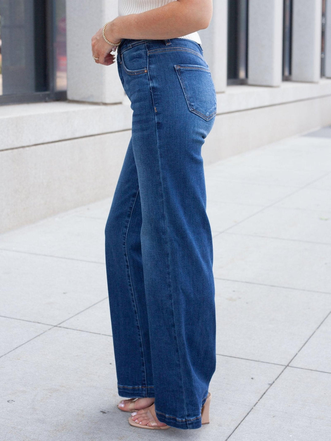 A person wearing blue wide-leg denim jeans, shown from the waist down on a light gray tiled sidewalk in front of a building.