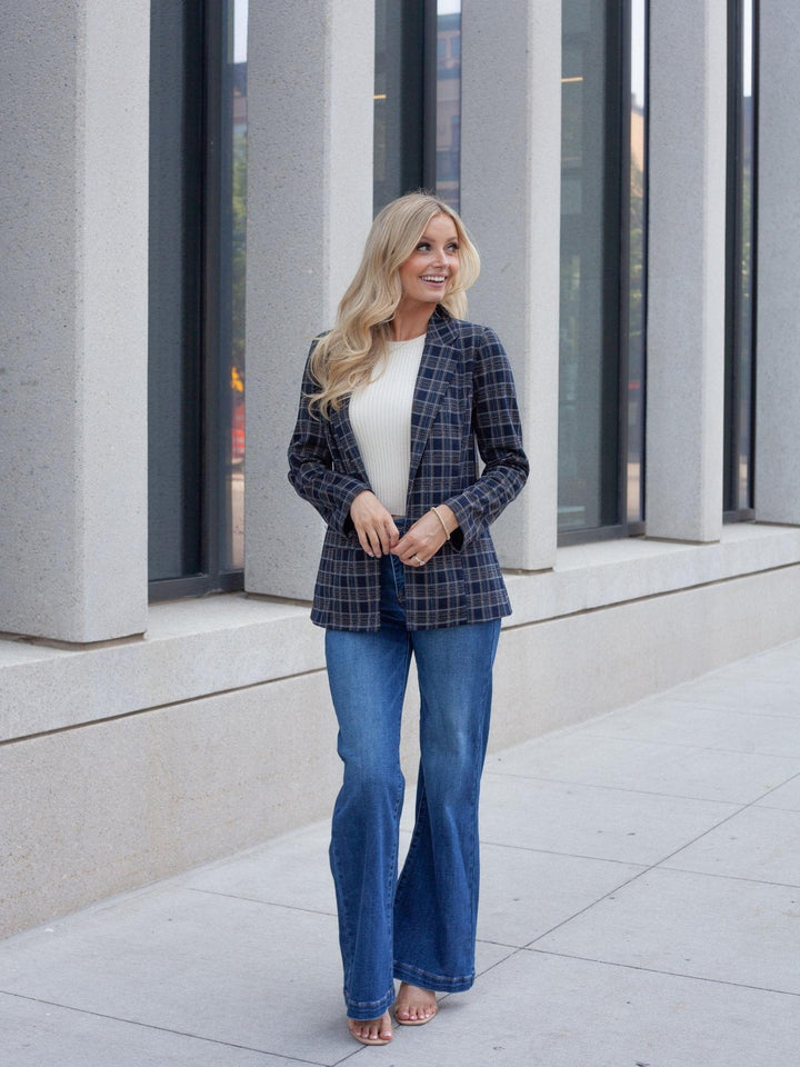 A woman stands outdoors on a sidewalk in front of a modern building with large gray columns and dark windows. She is wearing a navy and black plaid blazer over a white top, blue flare jeans, and is barefoot.