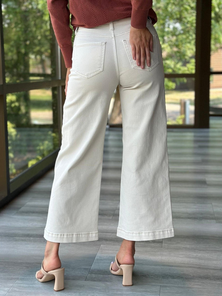 A person shown from the back wearing off-white cropped wide-leg jeans, a rust-colored knit top, and beige block-heel sandals in a modern indoor setting with large windows and a tiled floor.