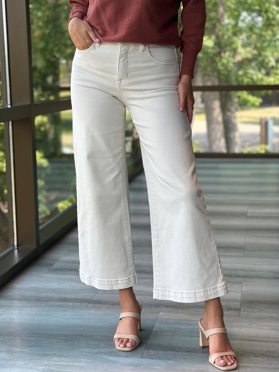 A person wearing off-white cropped wide-leg jeans with rolled hems, paired with a rust-colored long-sleeve top and beige heeled sandals, standing on a tiled balcony with a blurred outdoor background.