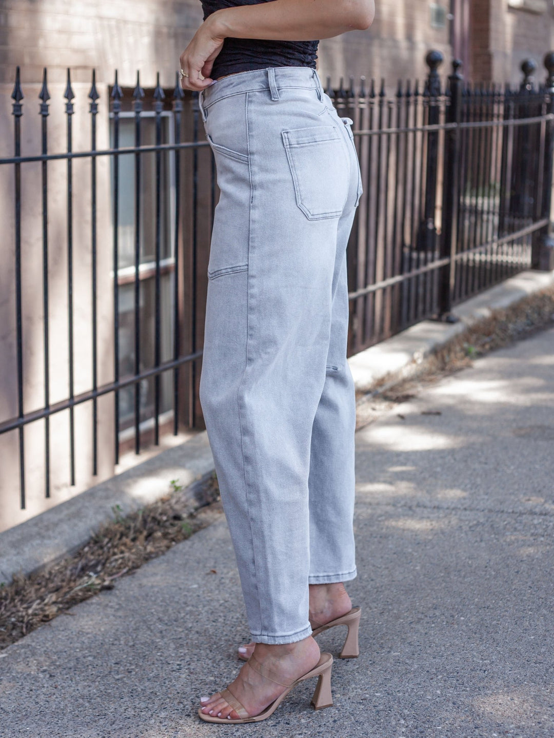 A person wearing light-wash, light-blue straight-leg denim jeans with a black top and clear-strap heeled sandals, standing on a sidewalk next to a black metal fence.