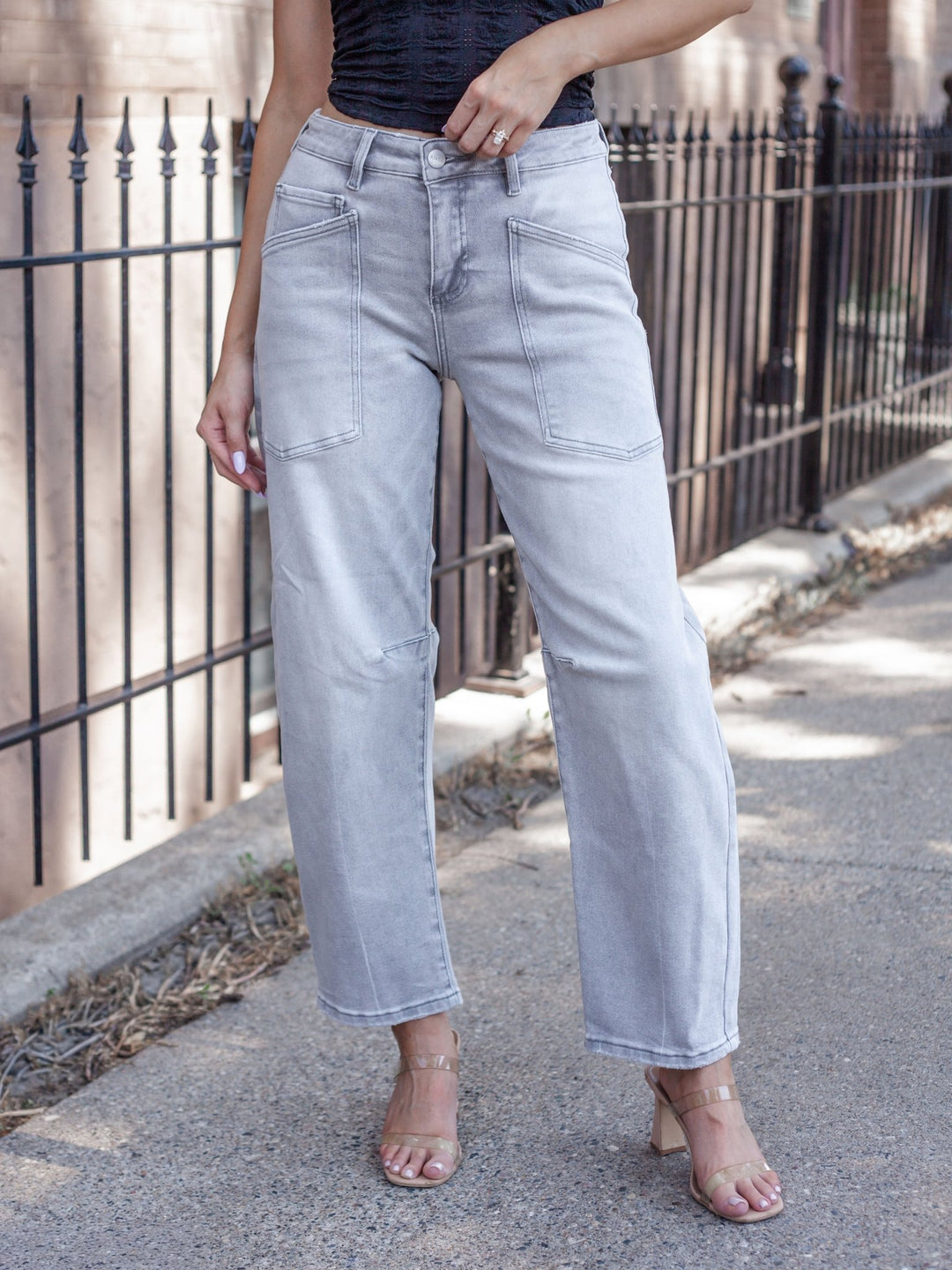 A person wearing light-wash gray cropped jeans with a black top and clear high-heeled sandals, standing on a sidewalk beside a black metal fence.