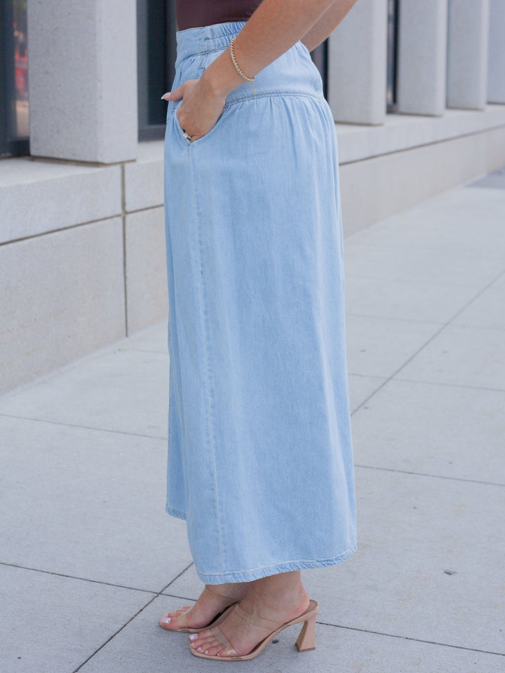 A person wearing a light blue denim maxi skirt with side pockets, paired with nude heeled mule sandals, standing outdoors on a paved walkway.