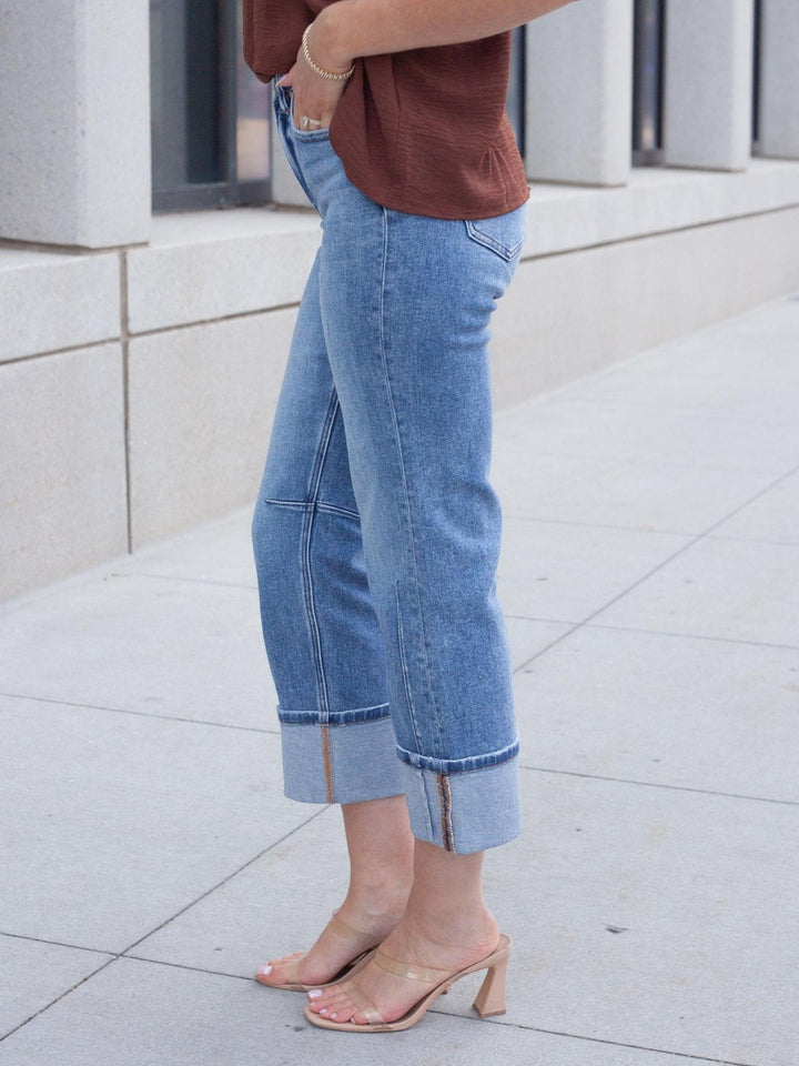A person wearing light blue cropped denim jeans with rolled cuffs and side zipper details, paired with a brown top and clear heeled sandals, standing on a light concrete sidewalk.