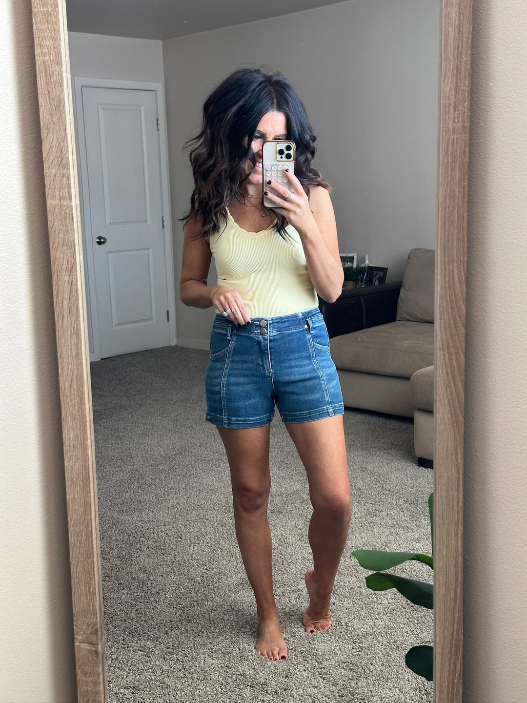 Person taking a mirror selfie wearing a yellow top and blue shorts in a room with a beige couch and white door.