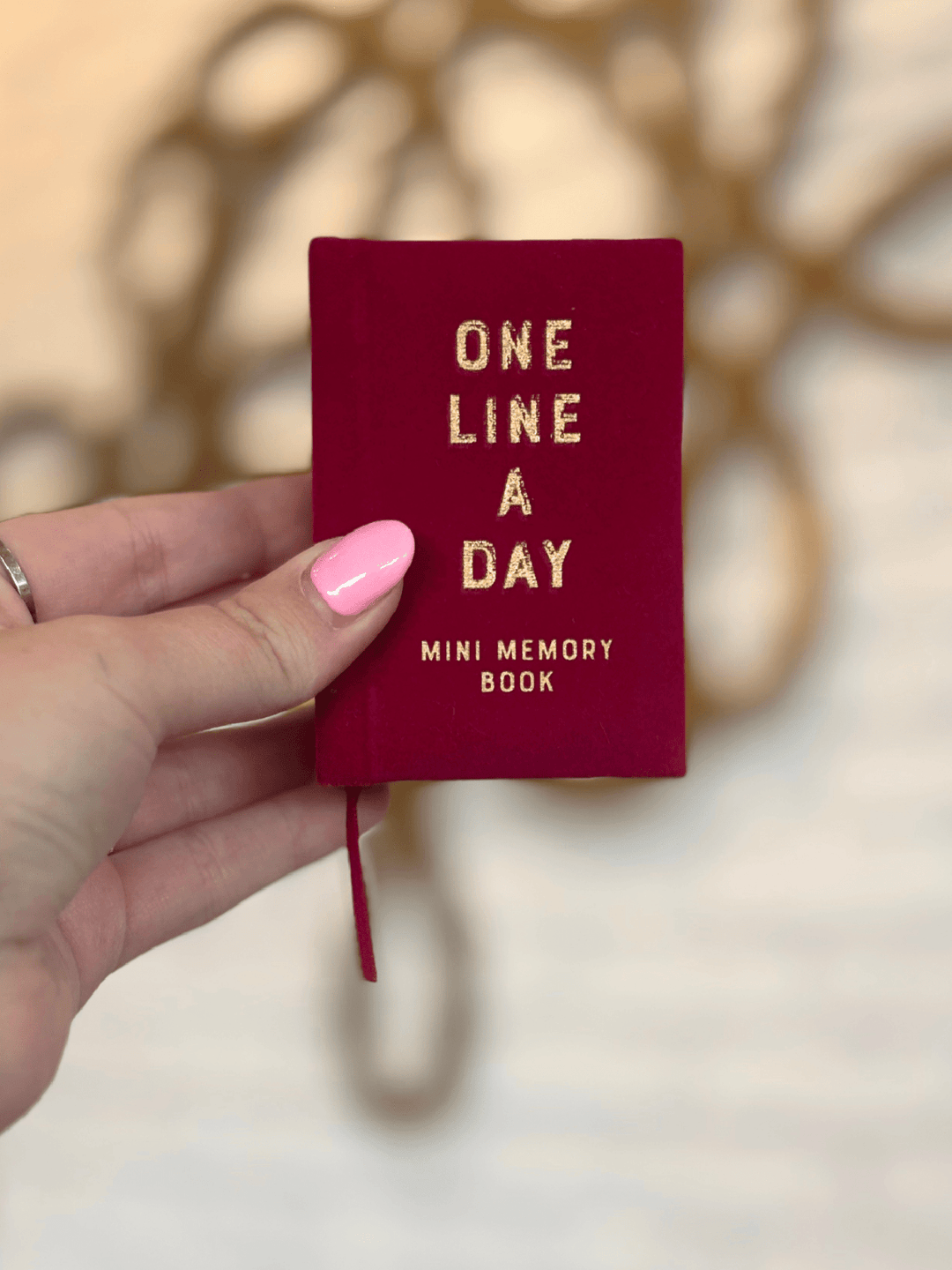 A hand with pink nail polish holds a small burgundy mini memory book with gold foil text that reads &