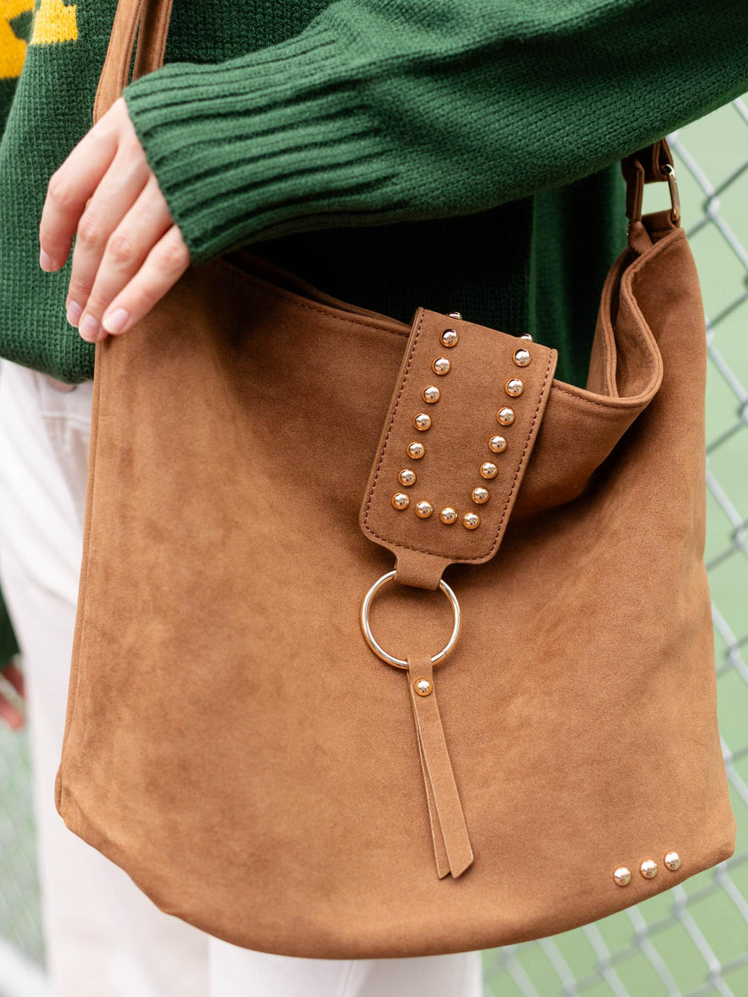 A tan suede shoulder bag with gold-tone studs and a circular ring detail, worn by a person in a green sweater.
