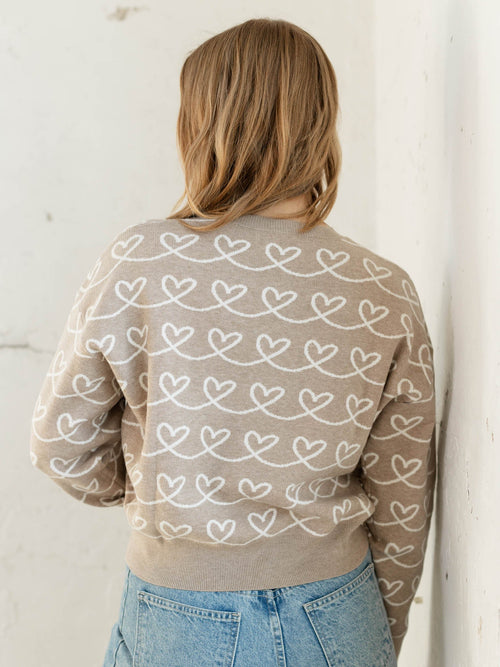 Person wearing a beige sweater with heart patterns against a white wall