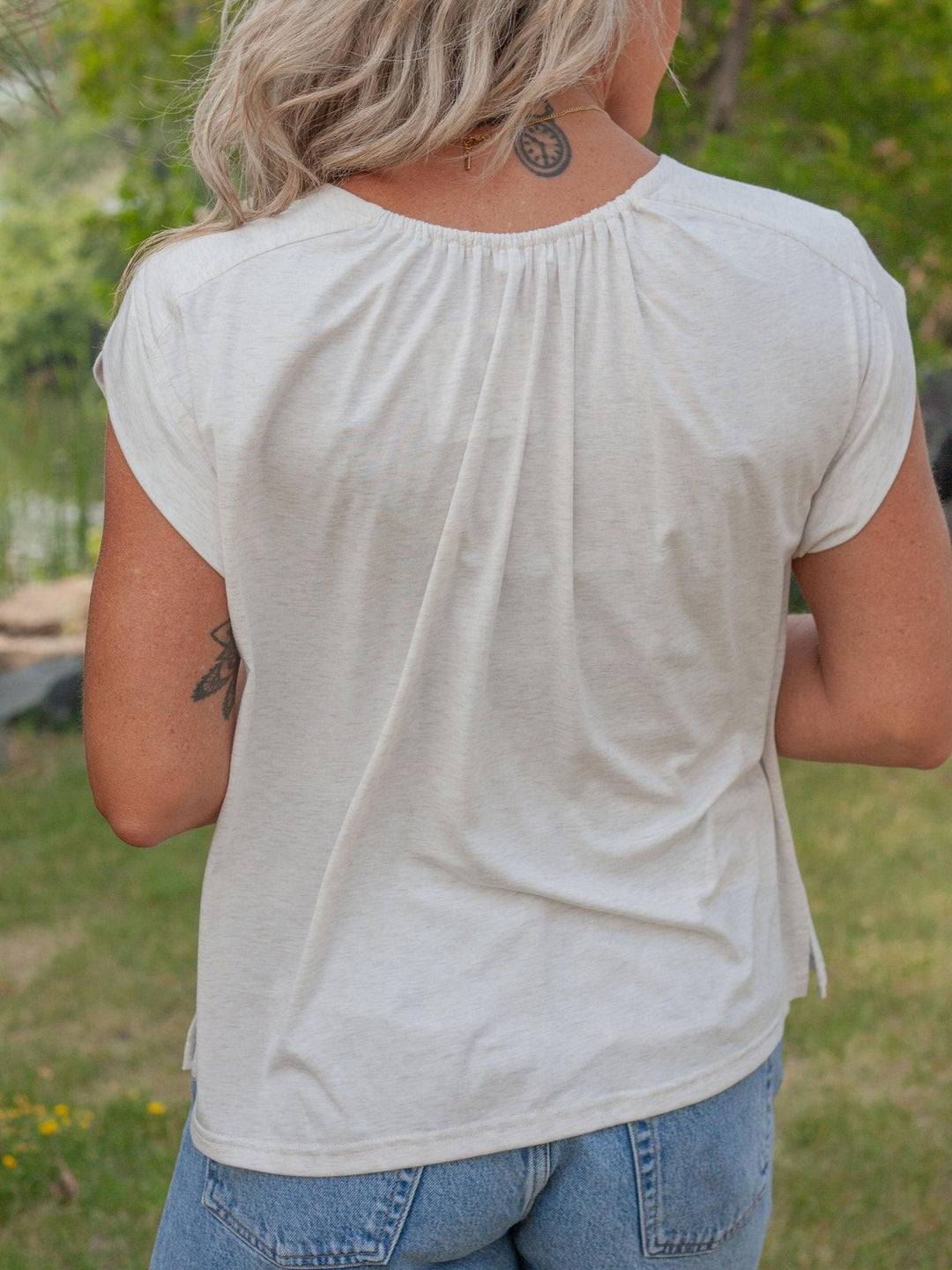 A woman viewed from behind wearing a light cream-colored short-sleeve top with a gathered neckline, paired with blue jeans, set against a blurred green outdoor background.