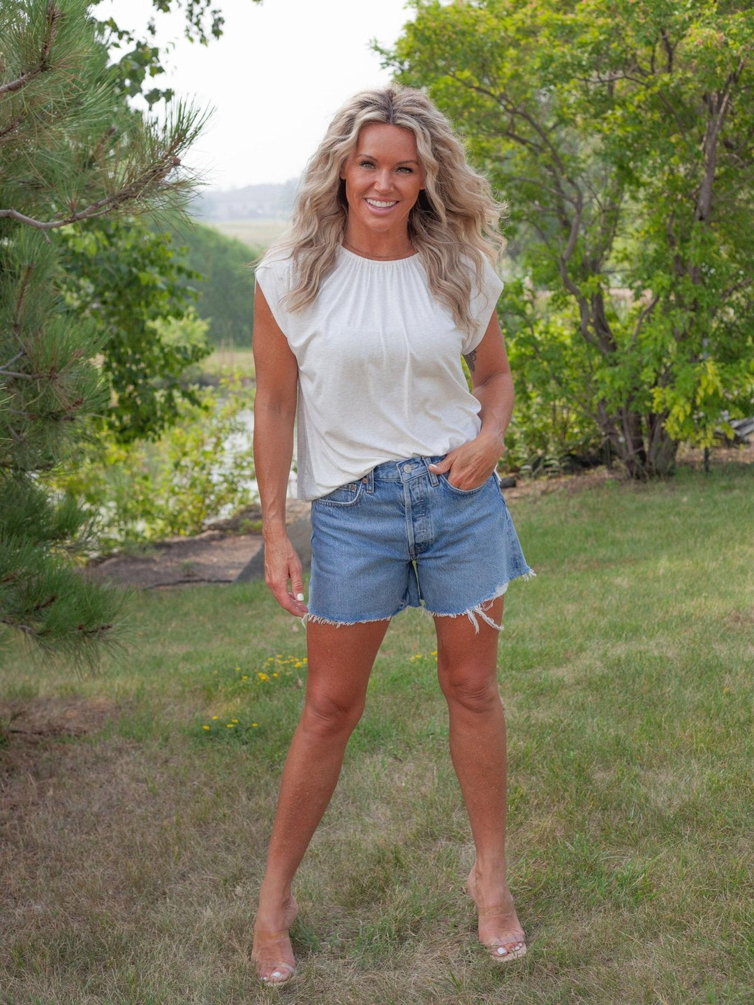 A woman standing outdoors on a grassy lawn wearing a white sleeveless top and light blue denim shorts, with trees and a body of water in the background.
