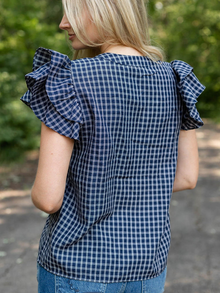 A woman shown from the back wearing a navy and white gingham check top with ruffled short sleeves, set against a blurred outdoor background of green foliage and a path.