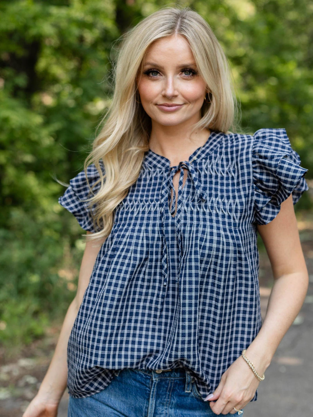 A woman with blonde hair wearing a navy and white gingham check blouse with ruffled short sleeves and a tie-neck detail, paired with blue jeans, standing outdoors against a blurred green foliage background.