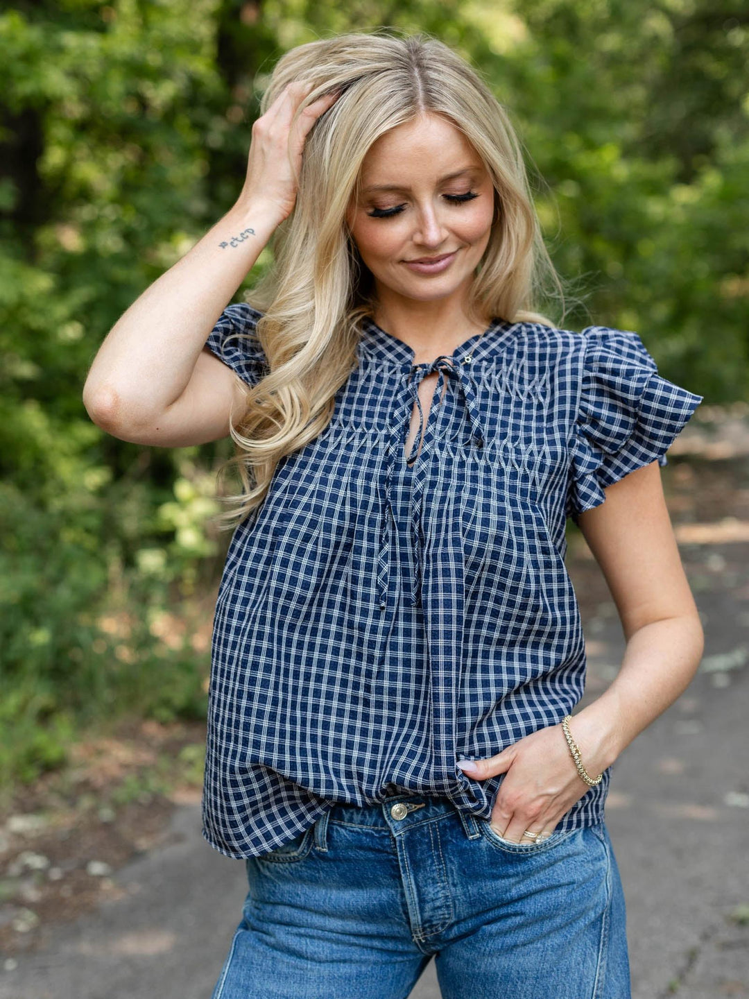 A woman wearing a navy and white gingham check short-sleeve blouse with ruffled shoulders and a tie-neck detail, paired with blue jeans, standing outdoors against a blurred green background.