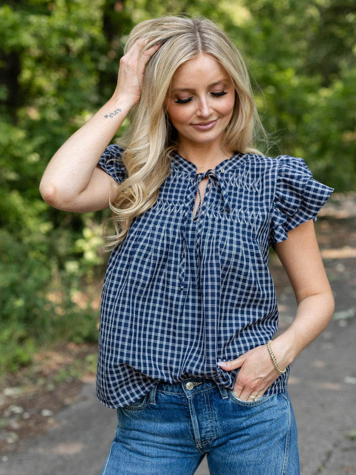 A woman wearing a navy and white gingham check short-sleeve blouse with ruffled shoulders and a tie-neck detail, paired with blue jeans, standing outdoors against a blurred green background.