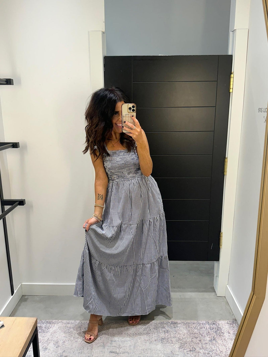 Woman taking a mirror selfie wearing a gray checkered dress in a room with a black wall.